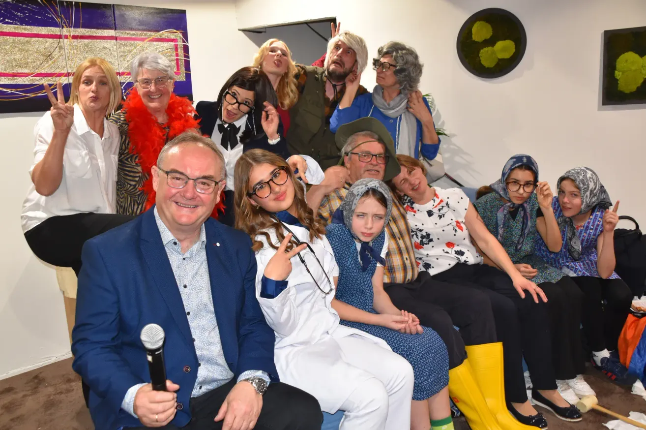 Eine Gruppe von Menschen in verschiedenen Kostümen sitzt zusammen auf einer Couch, lächelt und posiert für ein Foto. Ein Mann in einem blauen Anzug hält ein Mikrofon.