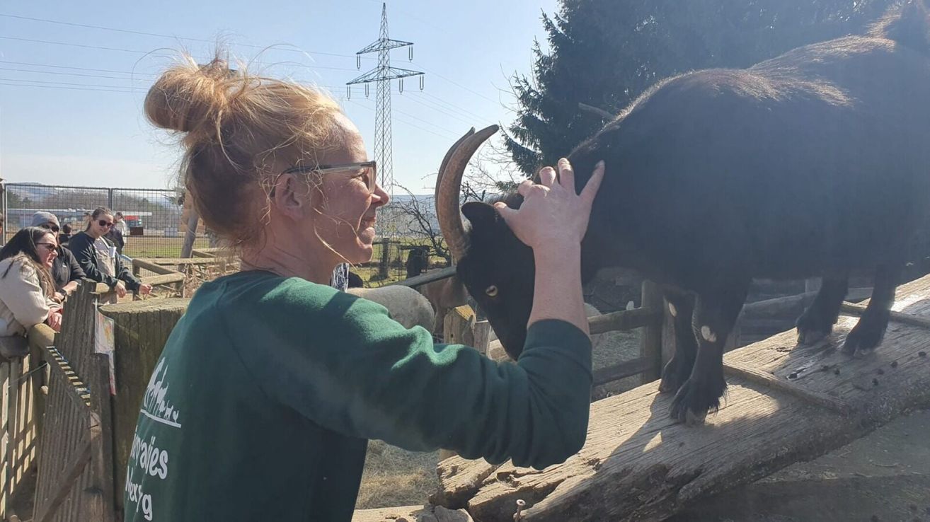 Eine Frau mit einer grünen Bluse und Brille streichelt einen schwarzen Ziegenbock. Der Bock hat große Hörner und weiße Flecken. Im Hintergrund befinden sich Bäume und ein Strommast.