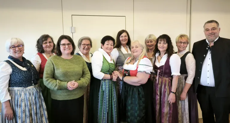 Eine Gruppe von Frauen, die lächeln, stehen eng beieinander in einem Raum. Die meisten von ihnen tragen traditionelle Kleidung. Sie stehen vor einer weißen Wand.