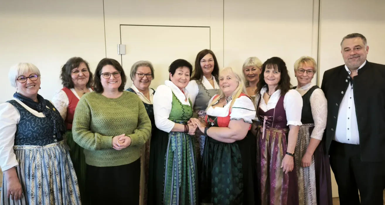 Eine Gruppe von Frauen, die lächeln, stehen eng beieinander in einem Raum. Die meisten von ihnen tragen traditionelle Kleidung. Sie stehen vor einer weißen Wand.