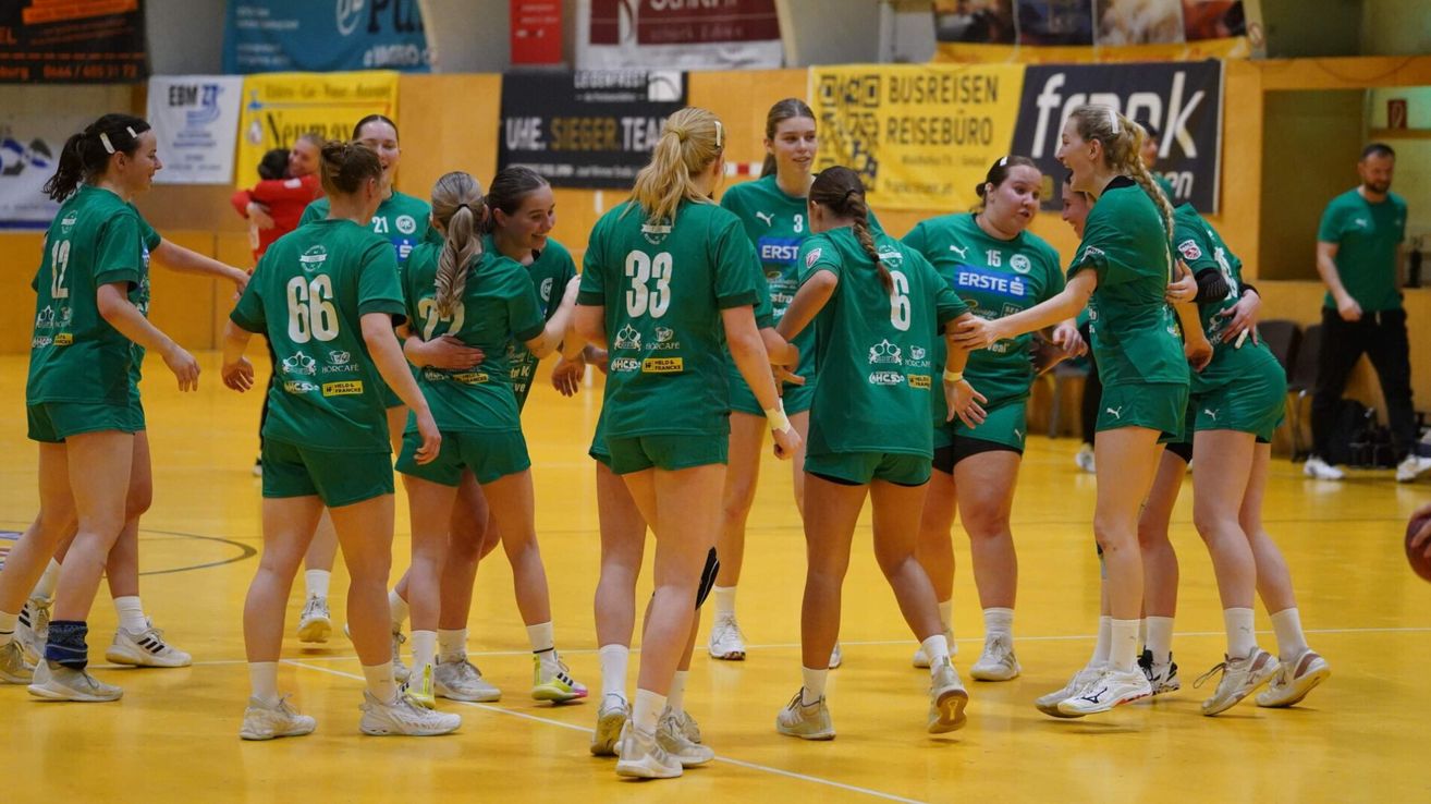 Eine Gruppe von Frauen in grünen Trikots und Turnschuhen feiert auf einem gelben Spielfeld, wobei die Nummern 33 und 6 auf ihren Uniformen sichtbar sind.