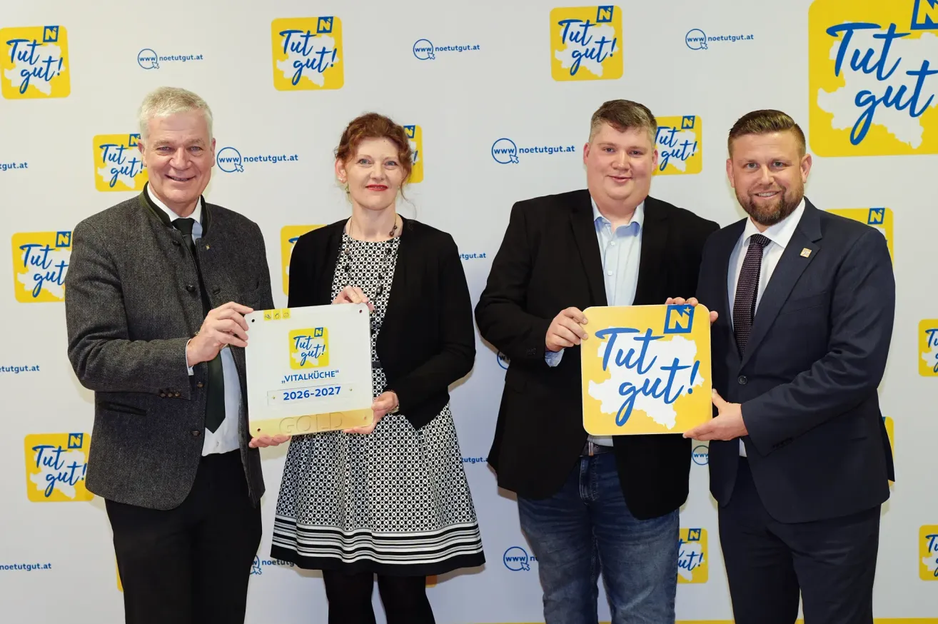 Vier Personen stehen vor einer Wand mit Auszeichnungen in der Hand. Die Wand hat gelbe und weiße Logos mit dem Text 'Tut gut!'. Die beiden Männer tragen Anzüge, während die beiden Frauen ein Kleid und eine Blazerjacke tragen. Sie alle lächeln und posieren für ein Foto.