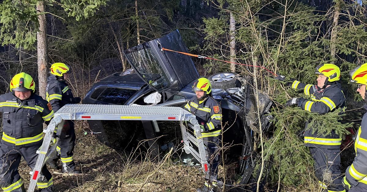 Feuerwehrleute arbeiten daran, ein beschädigtes Fahrzeug aus einem bewaldeten Gebiet zu extrahieren. Das Auto ist auf dem Dach mit offenem Motorraum, gesichert durch Seile.