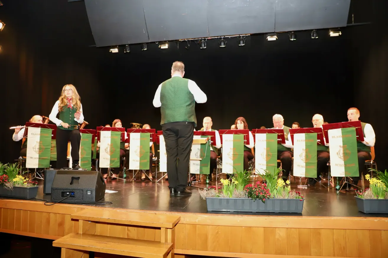 Eine Band tritt auf der Bühne auf, mit einem Mann davor. Dahinter sitzen andere Bandmitglieder und halten Musikinstrumente. Auf der Bühne befinden sich ein Lautsprecher und eine Blumenarrangement.