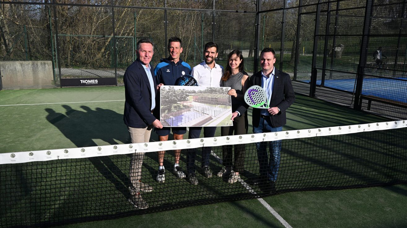 Fünf Personen stehen auf einem Tennisplatz und halten einen Schläger und ein Stück Papier. Sie lächeln, und die Frau hält einen Schläger in ihrer rechten Hand. Dahinter befindet sich ein Zaun mit einem Tor und Bäumen.