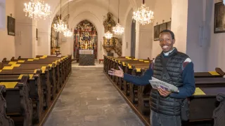 Ein Mann steht in einer Kirche, hält eine Zeitung und lächelt. Die Kirche hat hohe Decken mit Kronleuchtern, einen geschmückten Altar und Kirchenbänke.