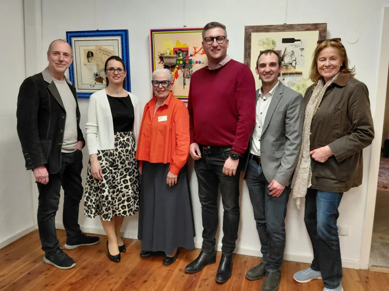 Sechs Personen posieren vor einer Wand mit verschiedenen gerahmten Kunstwerken und zeigen verschiedene Gesichtsausdrücke und Kleidung.