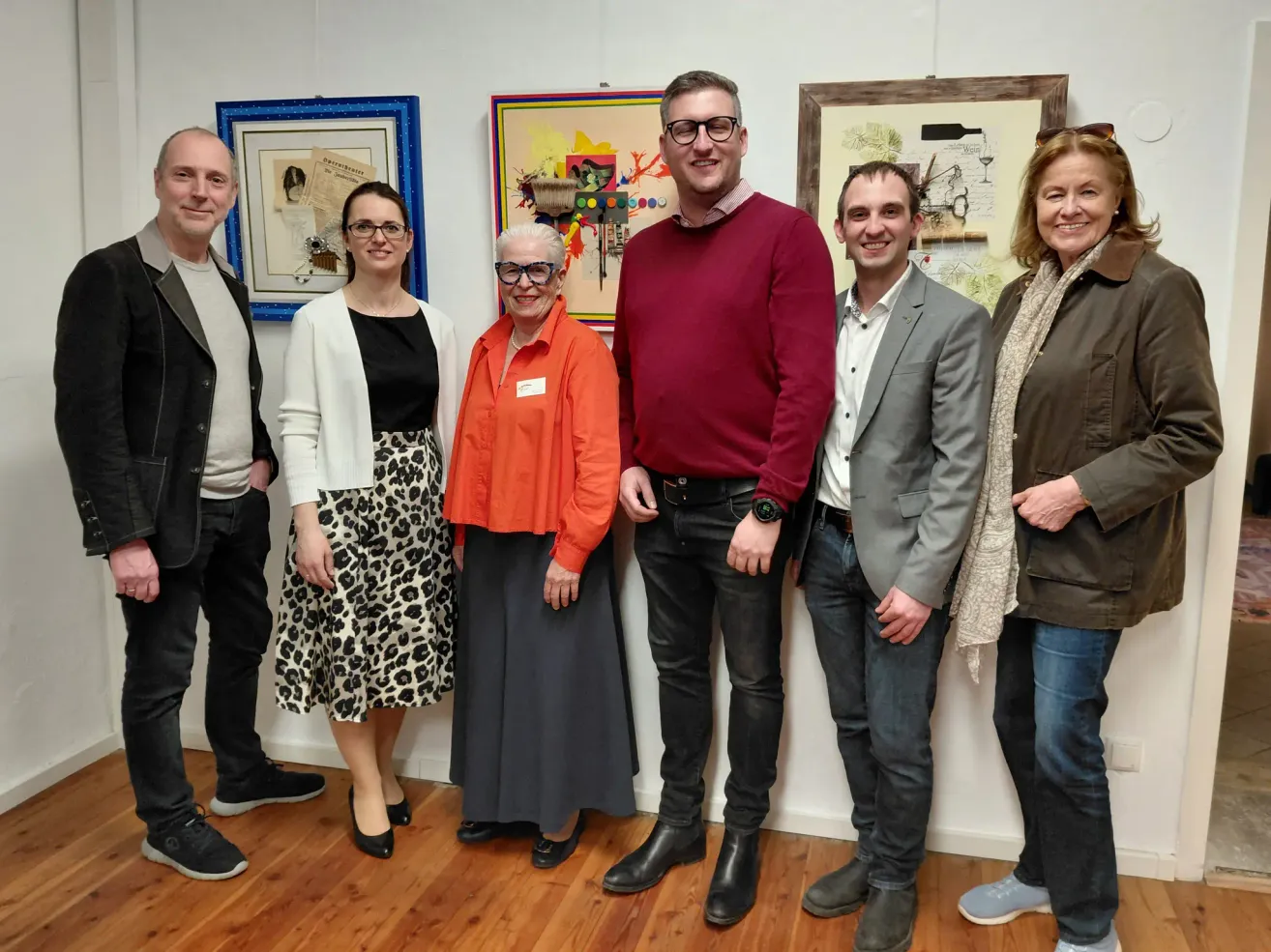 Sechs Personen posieren vor einer Wand mit verschiedenen gerahmten Kunstwerken und zeigen verschiedene Gesichtsausdrücke und Kleidung.