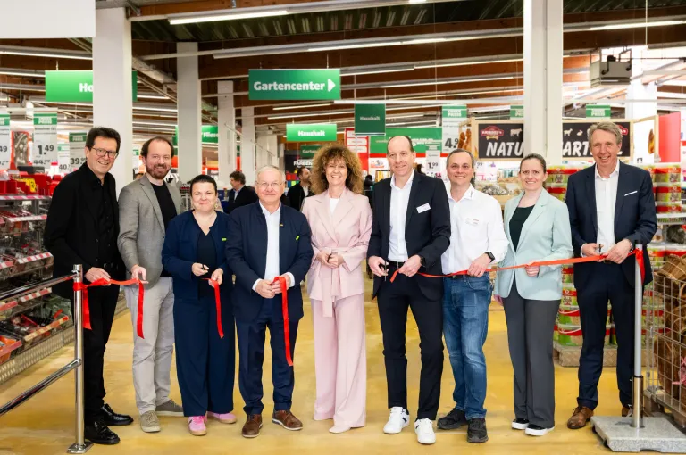 Eine Gruppe von Menschen steht in einem Geschäft zusammen und hält eine rote Schleife, wahrscheinlich zur Feier einer Eröffnung. Das Geschäft hat Schilder für Gartencenter und Haushalt.