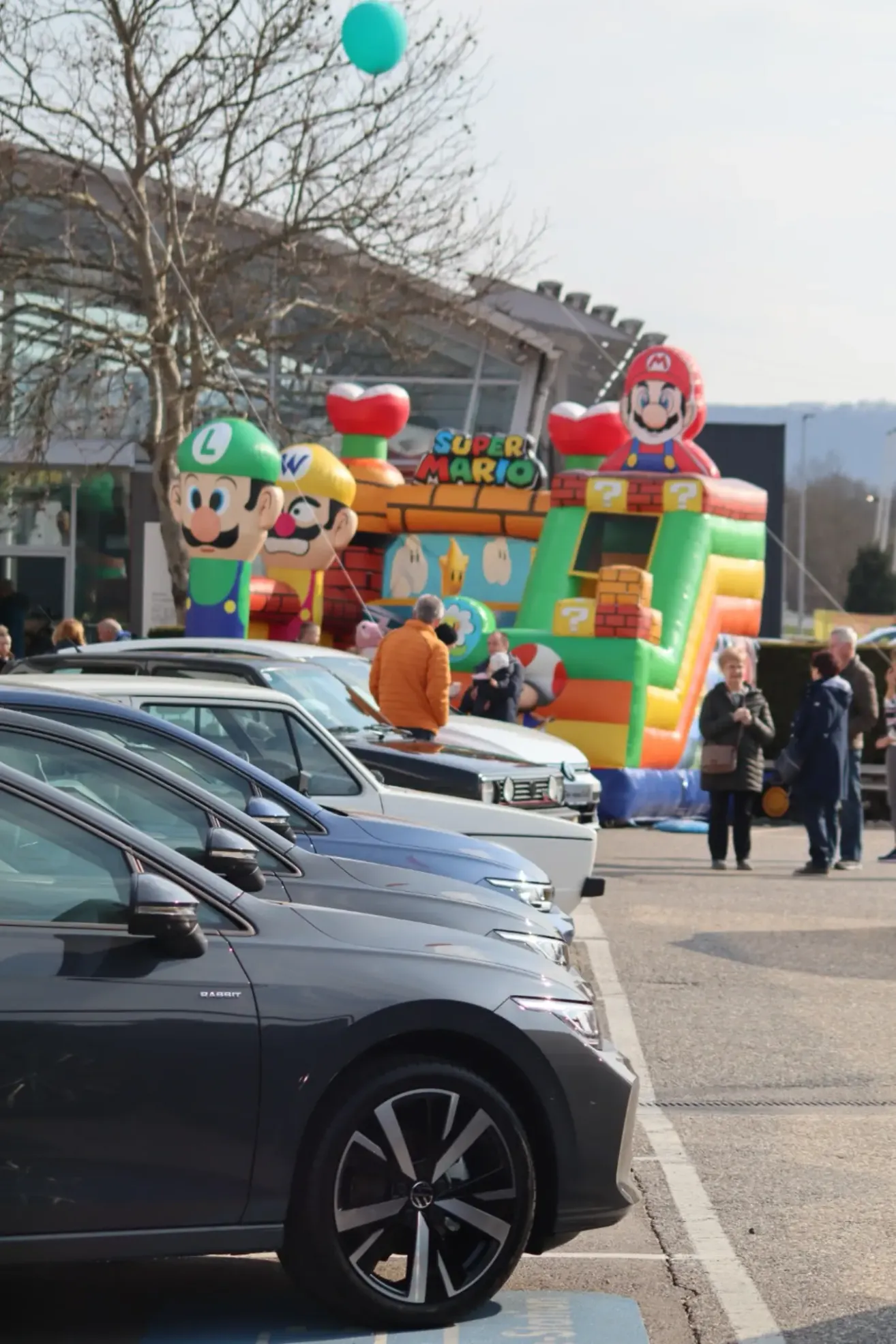 Ein Parkplatz mit Autos und einem großen Super Mario-Aufblasbaren. Leute gehen herum, einige stehen in der Nähe des Aufblasbaren.