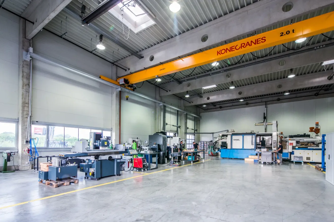 Ein Industriebetrieb mit verschiedenen Maschinen und Werkzeugen auf dem Boden. Ein großer gelber Kran ist von der Decke aufgehängt. Arbeiter bedienen einige der Maschinen.