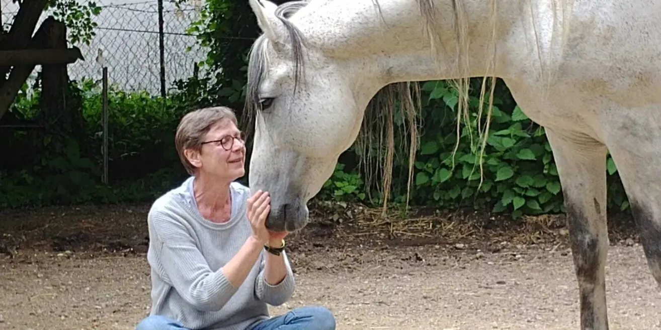 Eine ältere Frau mit Brille sitzt auf dem Boden und berührt das Gesicht eines weißen Pferdes. Das Pferd hat lange, wallende Mähne und Schweif. Sie sind von grünen Pflanzen und Erde umgeben.