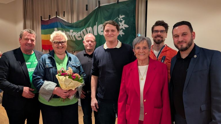 Fünf Menschen stehen lächelnd vor einer Regenbogenfahne. Eine Frau hält Blumen, und eine Fahne steht mit 'freunde' geschrieben.