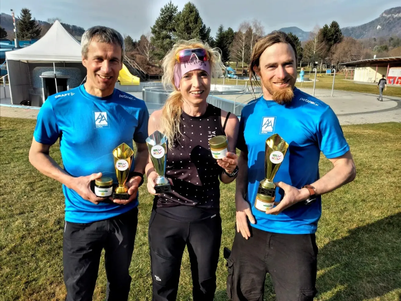 Drei Menschen in blauen Shirts posieren mit Trophäen für ein Foto. Eine Frau in einem schwarzen Top steht in der Mitte, und zwei Männer stehen auf beiden Seiten von ihr. Sie lächeln und halten Trophäen. Dahinter befindet sich ein grasbewachsenes Feld mit Bäumen und einem weißen Zelt.