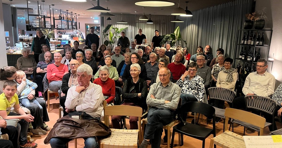 Ein Raum mit vielen Stühlen, die Menschen sitzen darin und reden. Ein Tisch mit Flaschen steht dahinter.