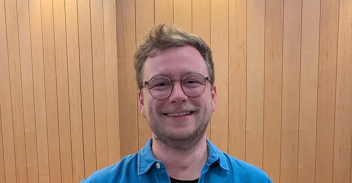 Ein Mann mit Bart und Brille lächelt für ein Foto vor einer Holzwand.