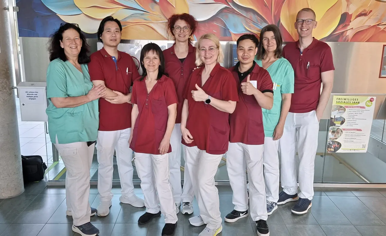 Eine Gruppe medizinischer Fachkräfte in roten Uniformen und weißen Hosen steht vor einer farbenfrohen Wandmalerei. Sie alle lächeln und posieren für ein Foto.