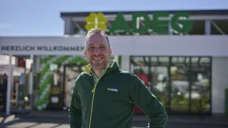 Ein Mann in einer grünen Jacke mit dem Wort ADE darauf steht vor einem Geschäft mit einem grünen Schild, auf dem ADE steht. Er lächelt in die Kamera.