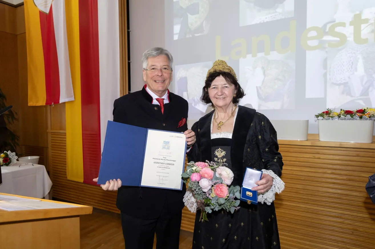 Ein Mann und eine Frau in traditioneller Kleidung halten ein Zertifikat und Blumen und stehen vor einer Flagge und einer Projektionsleinwand.
