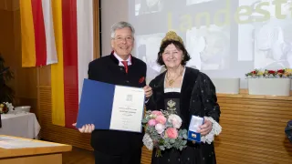 Ein Mann und eine Frau in traditioneller Kleidung halten ein Zertifikat und Blumen und stehen vor einer Flagge und einer Projektionsleinwand.