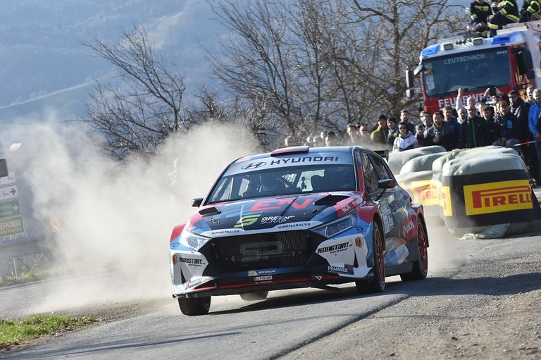 Ein Hyundai Rallye-Auto fährt auf einer staubigen Straße, mit Staubwolken hinter ihm. Zuschauer schauen von hinter Barrieren zu.