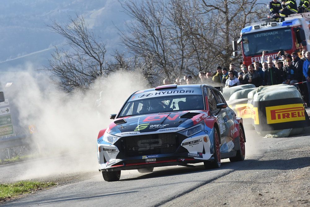 Ein Hyundai Rallye-Auto fährt auf einer staubigen Straße, mit Staubwolken hinter ihm. Zuschauer schauen von hinter Barrieren zu.