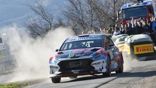 Ein Hyundai Rallye-Auto fährt auf einer staubigen Straße, mit Staubwolken hinter ihm. Zuschauer schauen von hinter Barrieren zu.