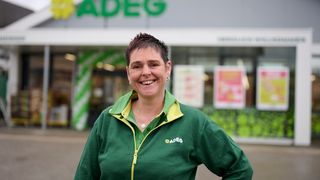 Eine Frau mit kurzen Haaren lächelt und posiert vor einem ADEG-Geschäft. Sie trägt eine grüne Jacke mit gelben Akzenten und eine Halskette. Hinter ihr ist das ADEG-Logo prominent am Gebäude angebracht.