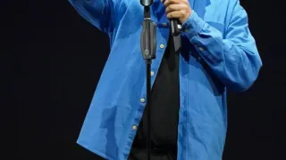 Ein Mann mit Brille und einem blauen Langarmhemd steht auf der Bühne und hält ein Mikrofon. Er lächelt und scheint eine Rede oder Präsentation zu halten.