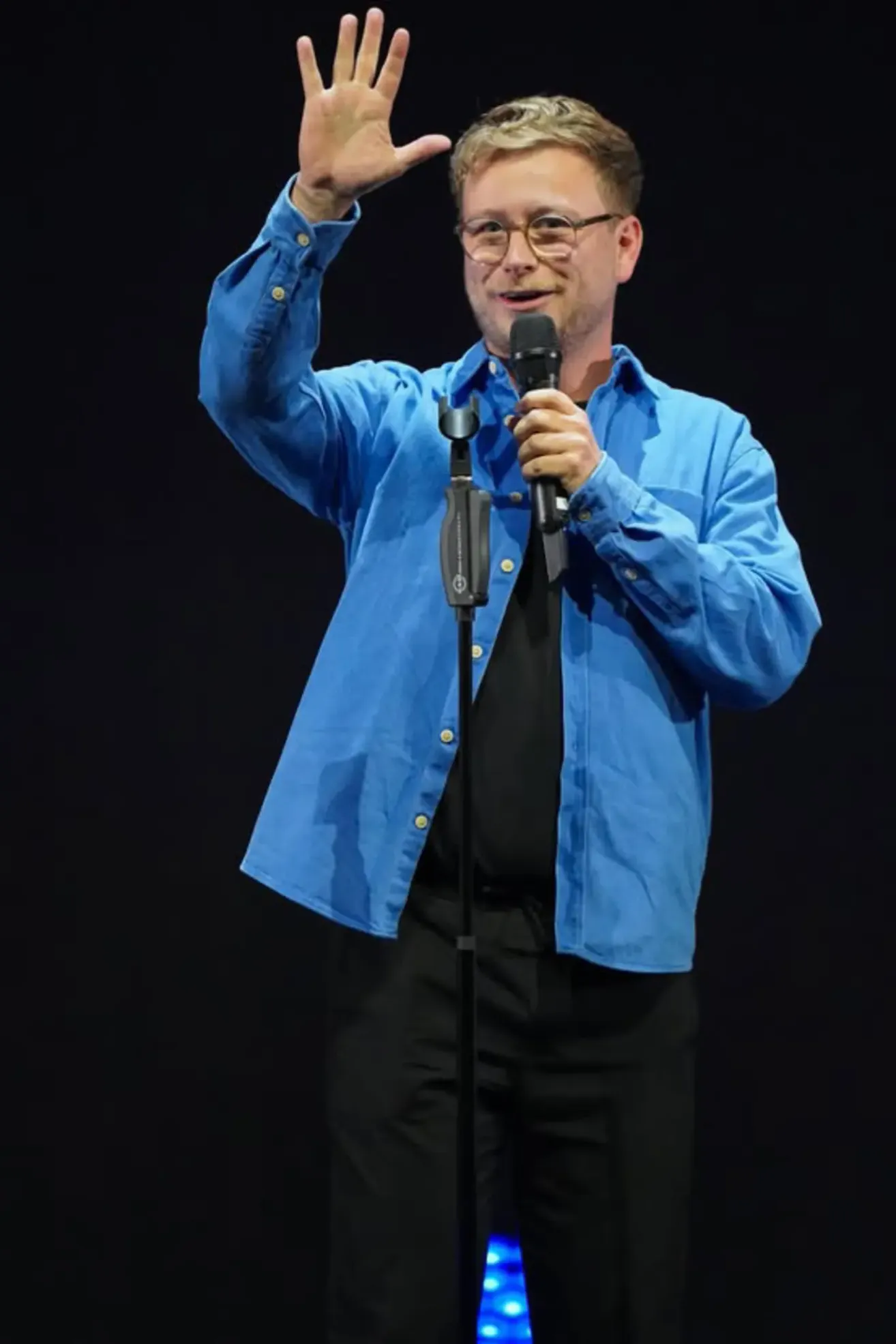 Ein Mann mit Brille und einem blauen Langarmhemd steht auf der Bühne und hält ein Mikrofon. Er lächelt und scheint eine Rede oder Präsentation zu halten.