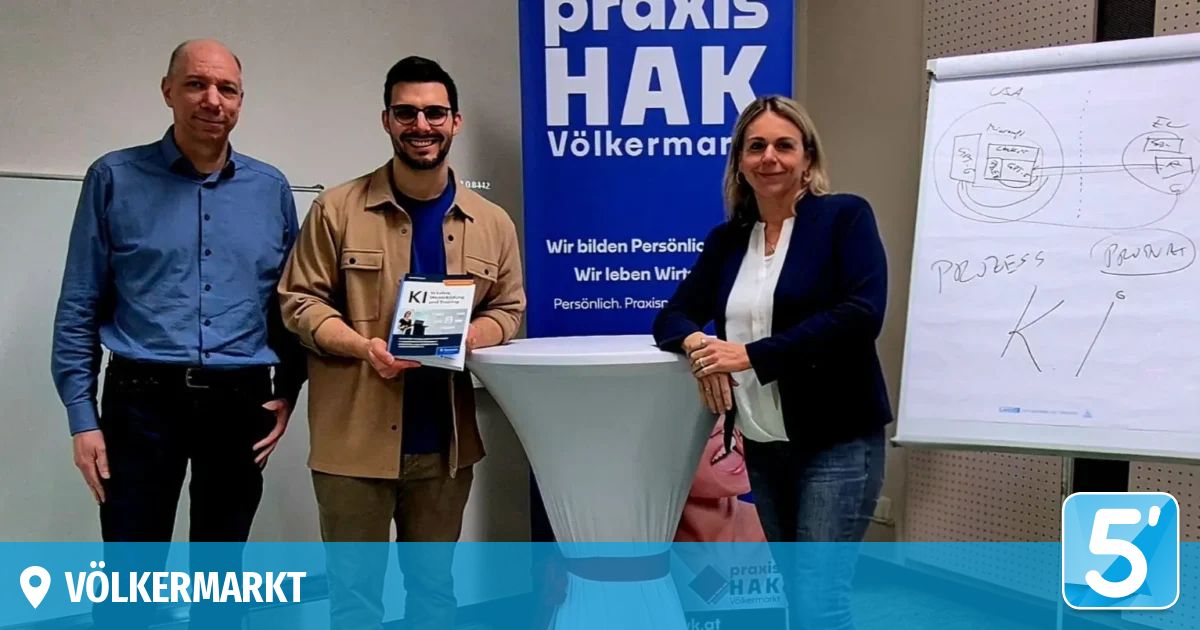 Drei Personen stehen vor einem weißen Tisch mit einem blauen Banner, auf dem 'Praxis HAK Volkermarkt' steht. Ein Mann in einem braunen Mantel hält ein Buch, während eine Frau in einem blauen Blazer in die Kamera lächelt. Dahinter ist ein Whiteboard teilweise zu sehen.
