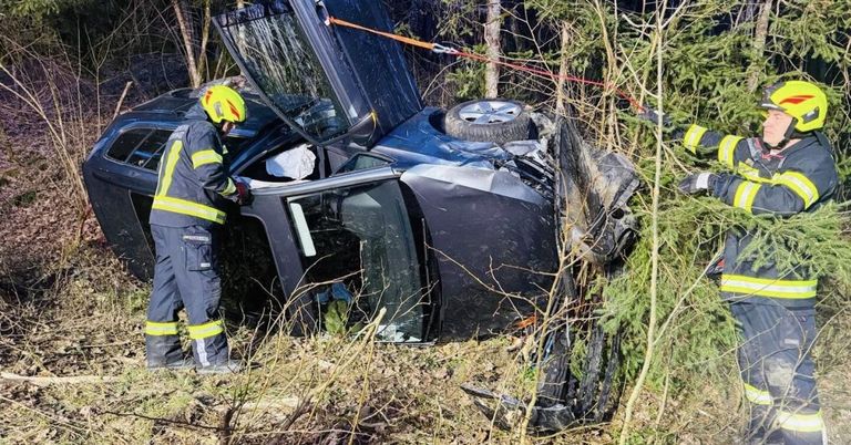 Ein Feuerwehrmann mit gelbem Helm inspiziert ein beschädigtes Fahrzeug, bei dem ein Reifen an der Seite hängt, in einem bewaldeten Gebiet.