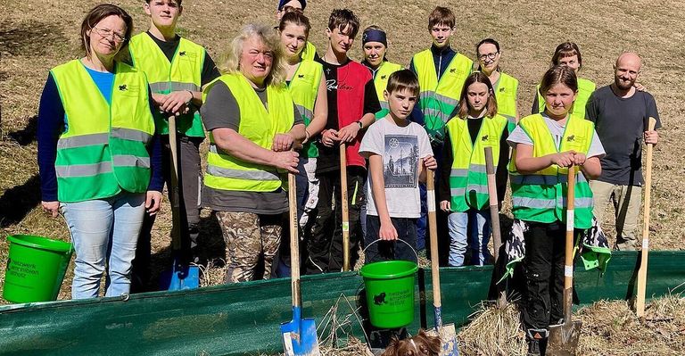 Eine Gruppe von Personen in Warnwesten, die im Freien stehen, Schaufeln in der Hand halten, mit einem grünen Eimer in der Mitte und einem Hund in der Nähe, vermutlich an einer Gemeinschaftsveranstaltung teilnehmend.