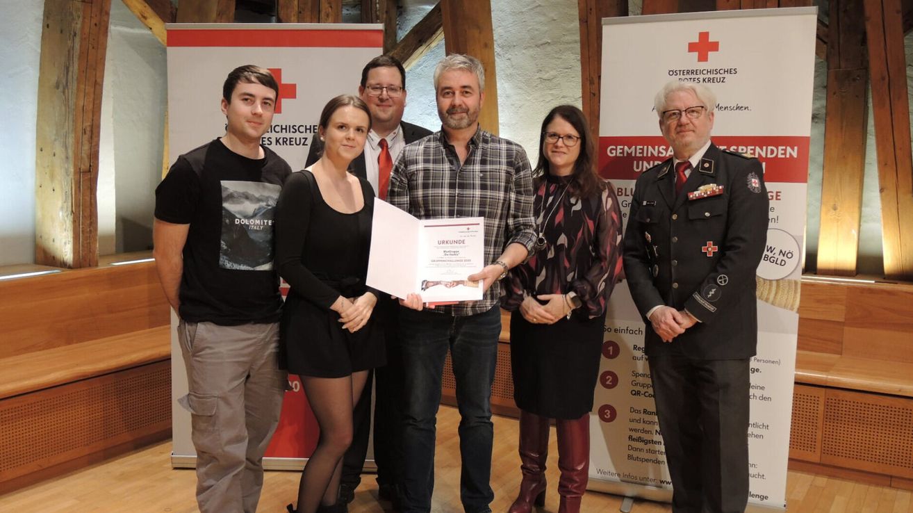 Eine Gruppe von Menschen steht zusammen und lächelt für ein Foto. Ein Mann in der Mitte hält ein Zertifikat, und zwei Frauen an seiner Seite tragen Brillen. Hinter ihnen sind Banner mit Text und Logos zu sehen.