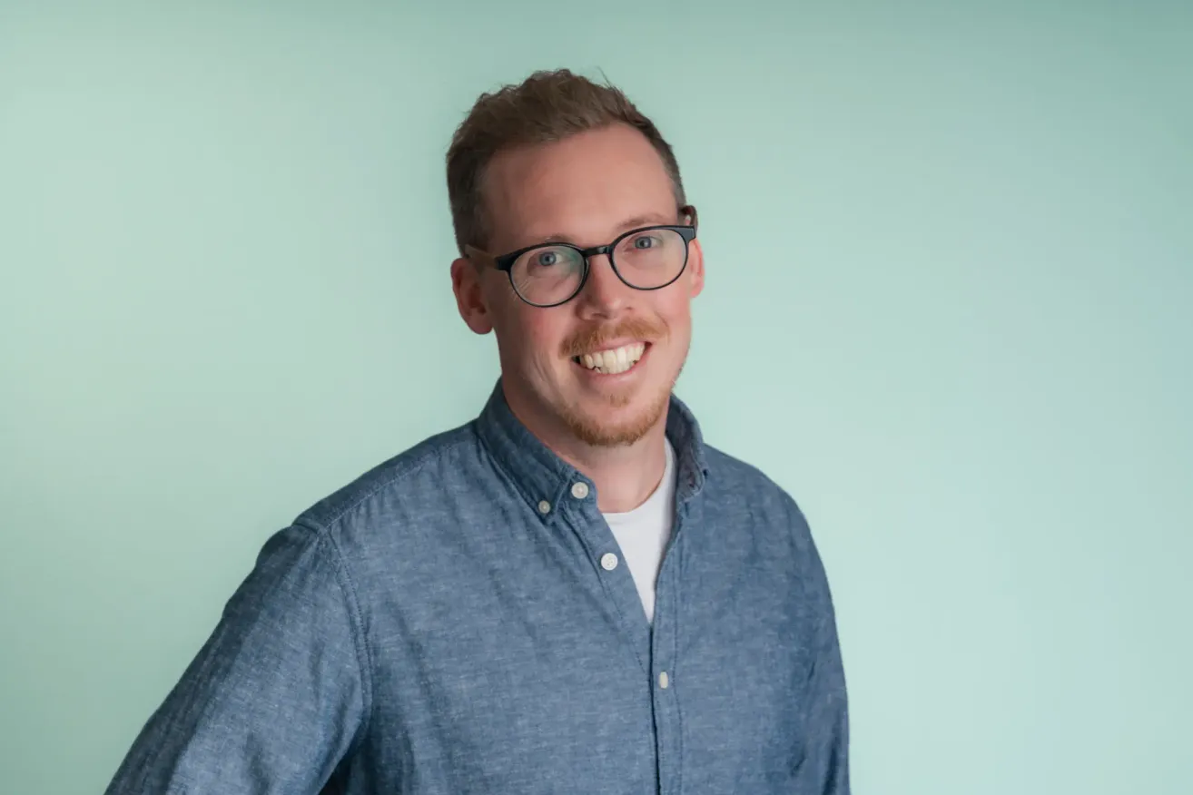Ein Mann mit Brille und Bart lächelt und posiert für ein Foto. Er trägt ein blaues Hemd mit weißem Kragen und hat einen hellgrünen Hintergrund.