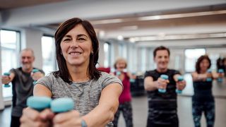 Eine Gruppe von Menschen in einem Fitnessstudio, die lächeln und mit blauen Hanteln trainieren. Eine Frau in der Mitte schaut direkt in die Kamera.