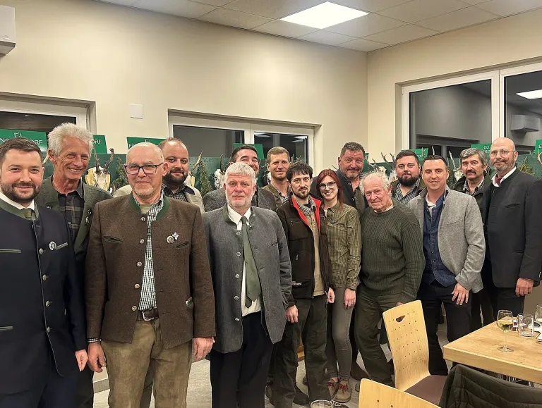 Eine Gruppe von Menschen steht drinnen und posiert für ein Foto. Sie sind in verschiedenen Kleidungsstücken gekleidet, und einige tragen Brillen. Hinter ihnen stehen Stühle und Tische. Der Raum hat weiße Wände, eine Deckenleuchte und Glasfenster.