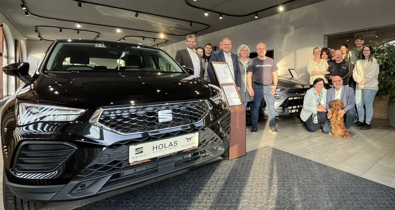 Eine Gruppe von Menschen posiert mit einem schwarzen Auto in einer Ausstellungshalle. Sie stehen hinter einem Plakat und in der Nähe eines Tisches. Das Auto ist ein SEAT HOLAS CUPRA mit einem Nummernschild.