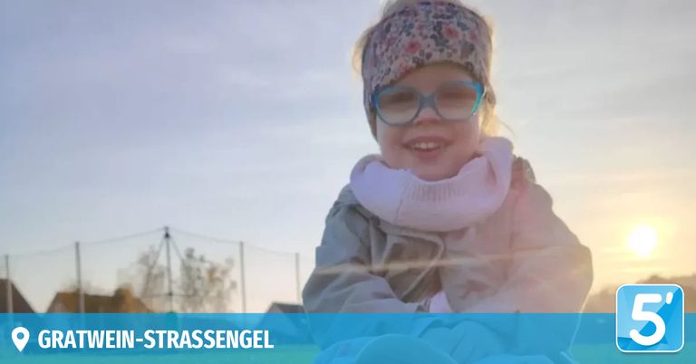 Ein junges Mädchen mit einer Brille, einem Blumenstirnband und einem rosa Schal lächelt im Freien. Der Hintergrund zeigt einen verschwommenen Zaun und Bäume. Das Wort 'STRASSEANGEL' ist unten sichtbar.