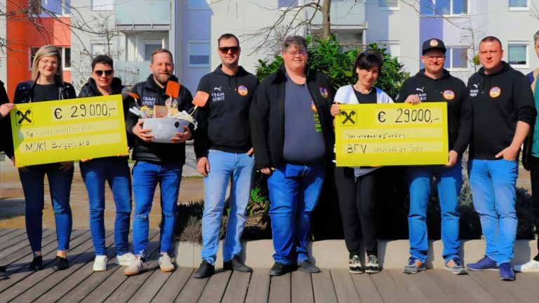 Fünf Personen stehen auf einer Holzplattform und lächeln für die Kamera. Eine hält eine Schüssel voller Gegenstände, während eine andere ein gelbes Schild mit dem Text € 2900 hält. Die Gruppe scheint eine Leistung zu feiern.