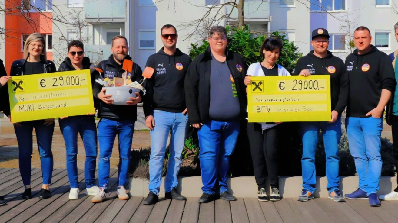 Fünf Personen stehen auf einer Holzplattform und lächeln für die Kamera. Eine hält eine Schüssel voller Gegenstände, während eine andere ein gelbes Schild mit dem Text € 2900 hält. Die Gruppe scheint eine Leistung zu feiern.