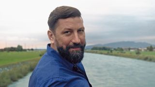 Ein Mann mit Bart und Schnurrbart in einem blauen Hemd steht am Fluss mit Bergen im Hintergrund.