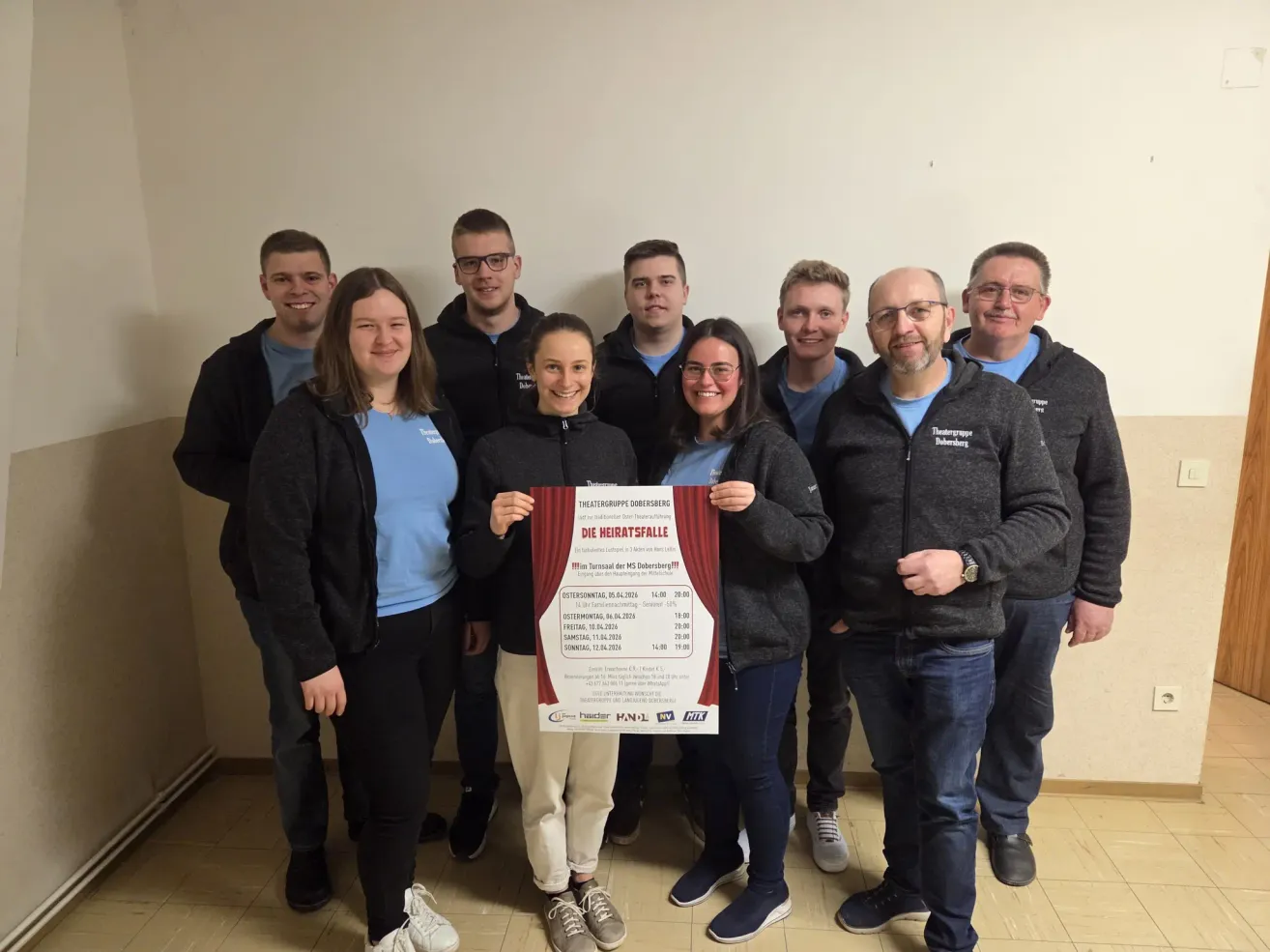 Eine Gruppe von Menschen steht zusammen für ein Foto, einige mit Brille und Jacken. Sie halten ein rotes Plakat mit weißem Text, betitelt 'Die Heiratsfalle.' Die Frau in der Mitte lächelt.