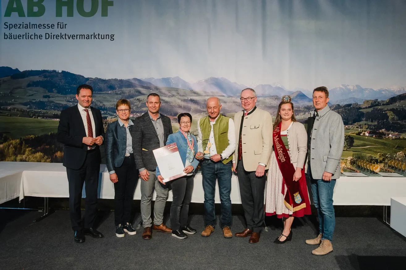 Eine Gruppe von Menschen in formeller Kleidung steht vor einem Hintergrund mit Bergen und einem Text, der 'Fovermarktung' lautet. Sie lächeln und scheinen für ein Foto zu posieren. Der Mann in der Mitte hält ein Zertifikat.