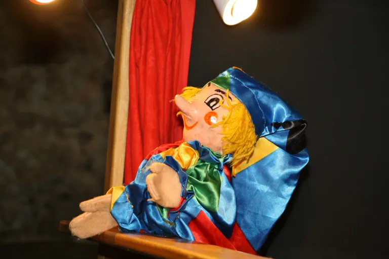 Eine farbenfrohe Puppe in blau, rot und gelb bekleidet sitzt auf einer Holzfläche in der Nähe eines roten Vorhangs. Der Kopf der Puppe ist geneigt und sie hat ein bemaltes Gesicht mit großen Augen und einer roten Nase.