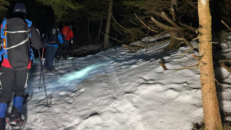Eine Gruppe von Wanderern mit Rucksäcken navigiert einen verschneiten Pfad bei Nacht, beleuchtet von einem grünen Licht. Sie sind von Bäumen und Schnee umgeben.