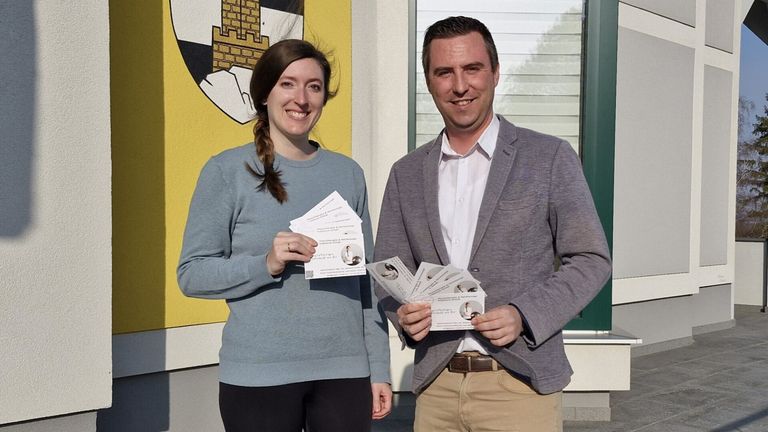 Ein Mann und eine Frau stehen lächelnd vor einem Gebäude und halten einen Stapel Visitenkarten in der Hand. Die Frau hat eine Zopf und trägt ein graues Shirt, während der Mann einen grauen Blazer trägt. Hinter ihnen ist eine gelbe Wand mit einem Wappen.