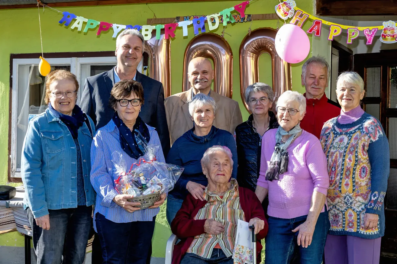 Gruppenfoto mit einer Hundertjährigen, ihrer Familie und Freunden bei einer Geburtstagsfeier. Sie stehen vor einem dekorierten Gebäude.