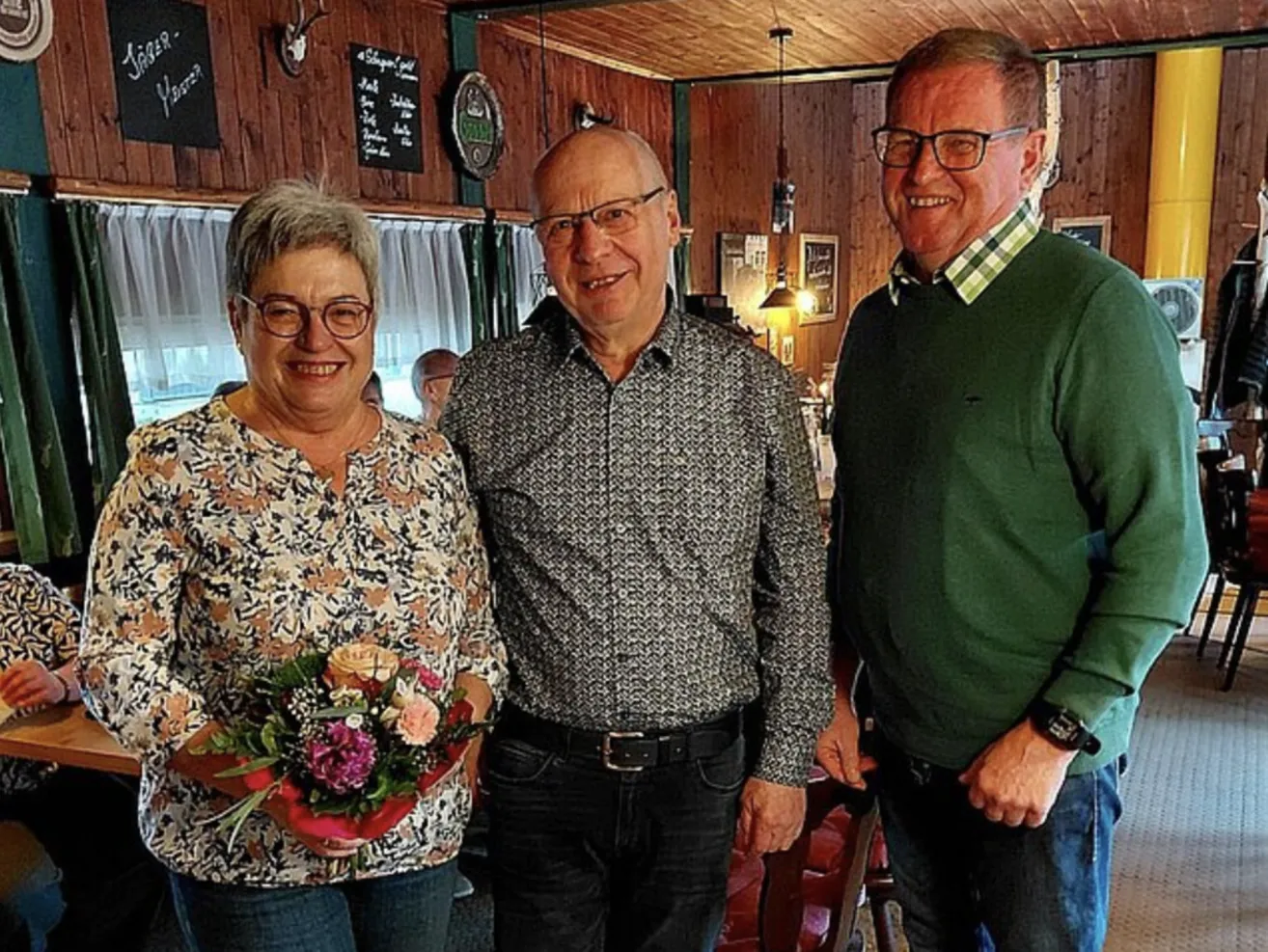 Drei Menschen stehen nebeneinander, lächelnd, in einem Raum mit Holzwänden. Eine Frau hält einen Blumenstrauß. Ein Mann auf der rechten Seite trägt einen grünen Pullover und eine Uhr. Ein Mann in der Mitte trägt ein graues Hemd und einen schwarzen Gürtel.