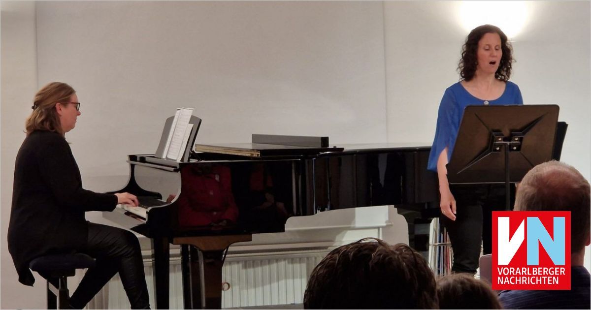 Ein Pianist spielt Klavier, während eine Frau singt. Einige Leute schauen zu. Der Raum ist mit warmer Beleuchtung erhellt.